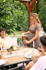 A pregnant woman presents a bottle of champagne at an outdoor summer dinner party with friends.

