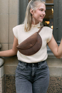 A blonde woman models a stylish brown leather half-moon crossbody bag with gray jeans and a cream sweater vest.
