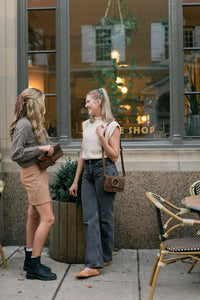 Two stylish women wearing fall fashion outfits chat outside a boutique shop with vintage-inspired leather handbags.
