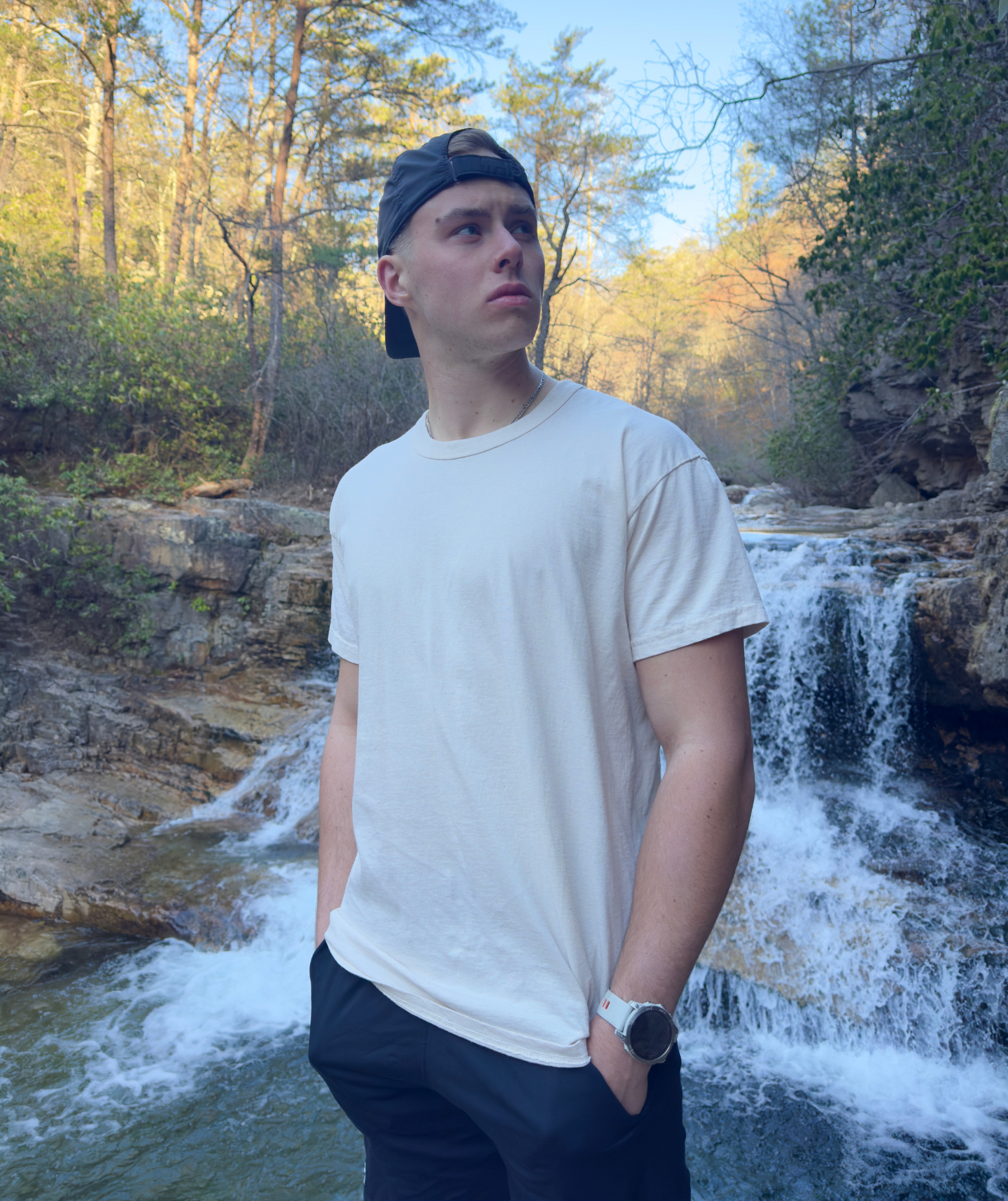 A young man wearing a baseball cap stands with his hands in his pockets, gazing at a cascading waterfall in a lush forest.
