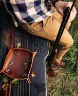 A hunter sitting on a truck tailgate holds his rifle beside a leather tray filled with ammunition.
