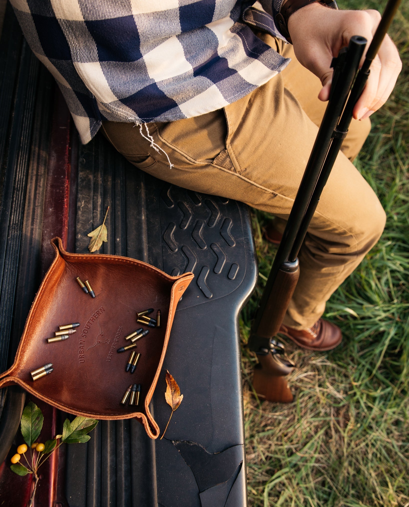 A hunter sitting on a truck tailgate holds his rifle beside a leather tray filled with ammunition.
