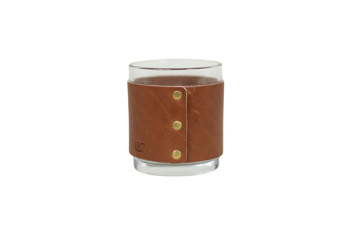 Close-up shows a clear glass tumbler wrapped in a brown leather sleeve with brass button details and an embossed "US" logo, set against a white background.
