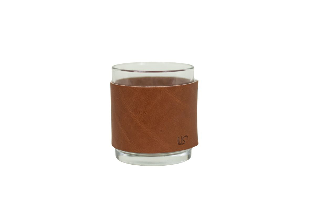 A clear glass tumbler wears a stylish brown leather sleeve with subtle embossed initials.

