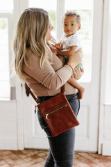 A woman carries her toddler son while wearing a stylish brown leather crossbody bag with a zipper pocket.
