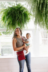 A mother smiles while holding her baby indoors amidst hanging ferns and wearing a red crossbody purse.
