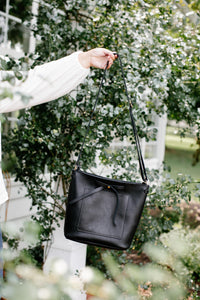A person holds up a stylish black leather crossbody bag outdoors.
