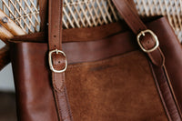 A brown leather tote bag with gold buckles sits on a wicker chair.

