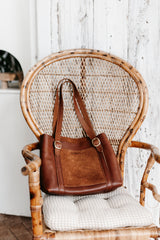 A brown leather tote bag rests on a wicker peacock chair against a rustic white wood backdrop.

