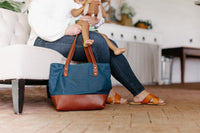 A woman holds her child with a stuffed animal while a blue canvas and brown leather tote bag rests at her feet.
