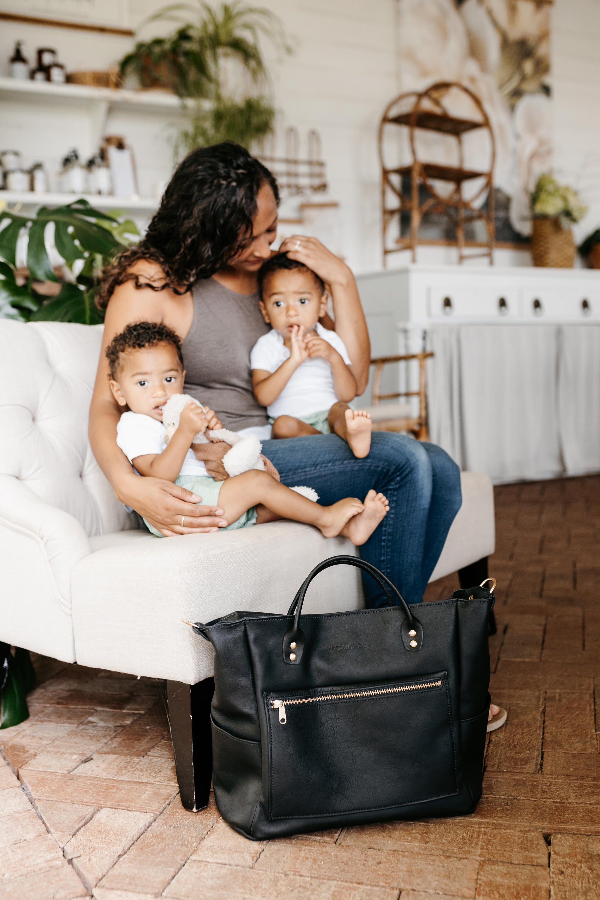 A mother lovingly cradles her two adorable biracial baby sons on a tufted chair, with a stylish black diaper bag nearby.
