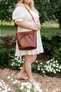 A woman wearing a white eyelet dress showcases a stylish brown leather tote bag while barefoot among white flowers in a garden setting.

