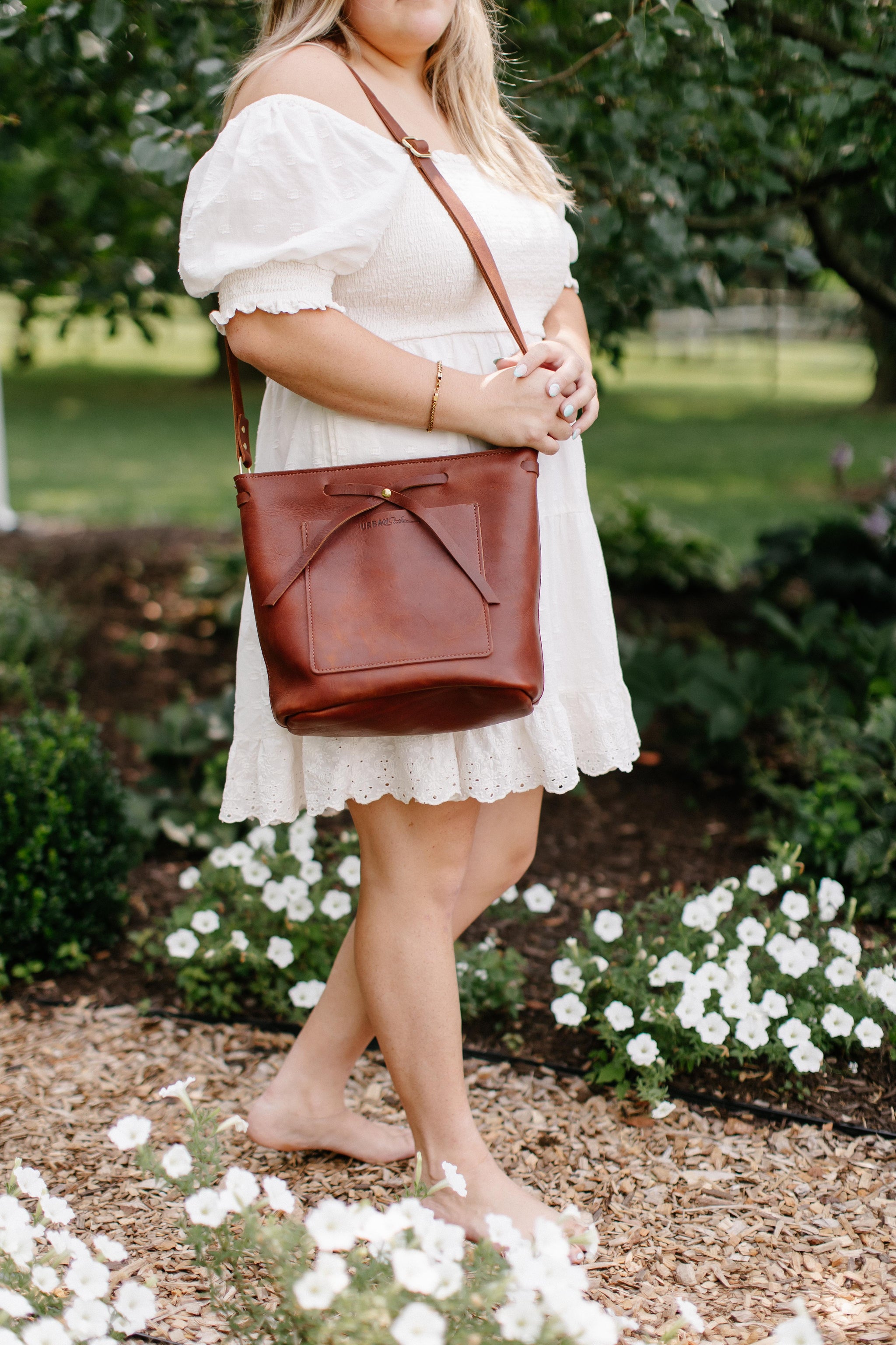 A woman wearing a white eyelet dress showcases a stylish brown leather tote bag while barefoot among white flowers in a garden setting.
