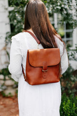 Woman carries a handcrafted, brown leather backpack while wearing a white dress outdoors.
