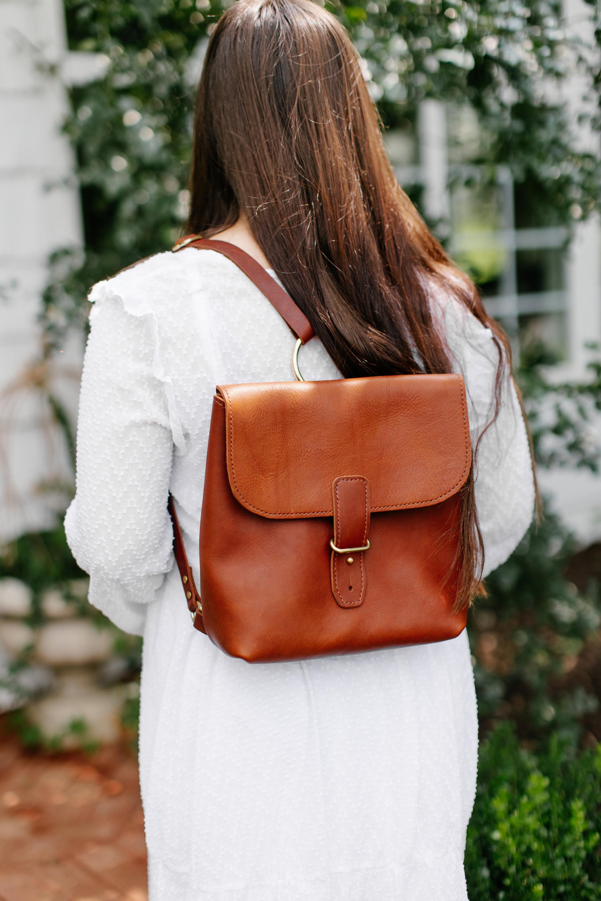 Woman carries a handcrafted, brown leather backpack while wearing a white dress outdoors.
