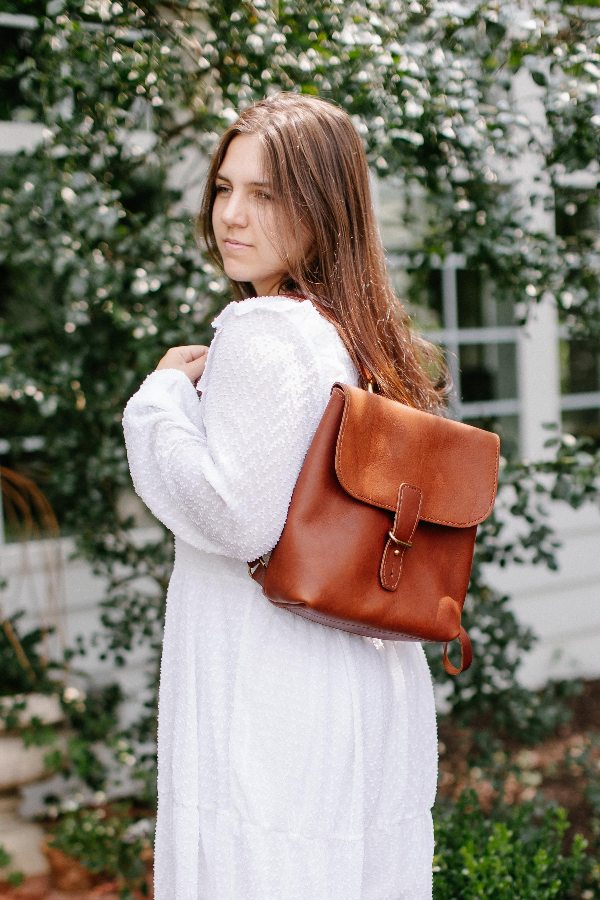 Woman in white dress models a brown leather backpack outdoors.
