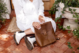 Woman sits on a brick patio with plants, showcasing a stylish brown leather backpack and white ankle boots.
