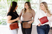 Three smiling women model stylish leather crossbody bags in a bright, plant-filled studio.
