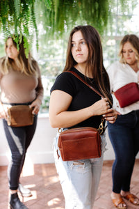 A young woman models a brown leather crossbody bag with a gold chain strap in a bright greenhouse setting, showcasing the stylish design.
