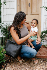 A smiling mother, wearing a stylish leather handbag, lovingly holds her young toddler outside their home.
