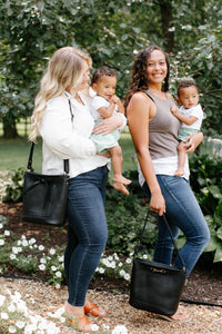 Two smiling women each carry a baby while showcasing stylish black leather bucket bags in a garden setting.

