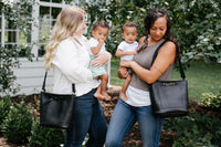 Two mothers each carry a toddler while showcasing stylish black leather tote bags outdoors.
