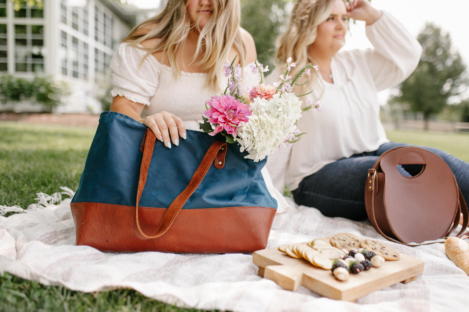 Two stylish women enjoy a blissful picnic with flowers, artisan cheese board, and fashionable tote bags on a sunny day outdoors.
