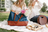 Two stylish women enjoy a blissful picnic with flowers, artisan cheese board, and fashionable tote bags on a sunny day outdoors.
