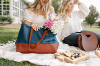 Two stylish women enjoy a blissful picnic with flowers, artisan cheese board, and fashionable tote bags on a sunny day outdoors.
