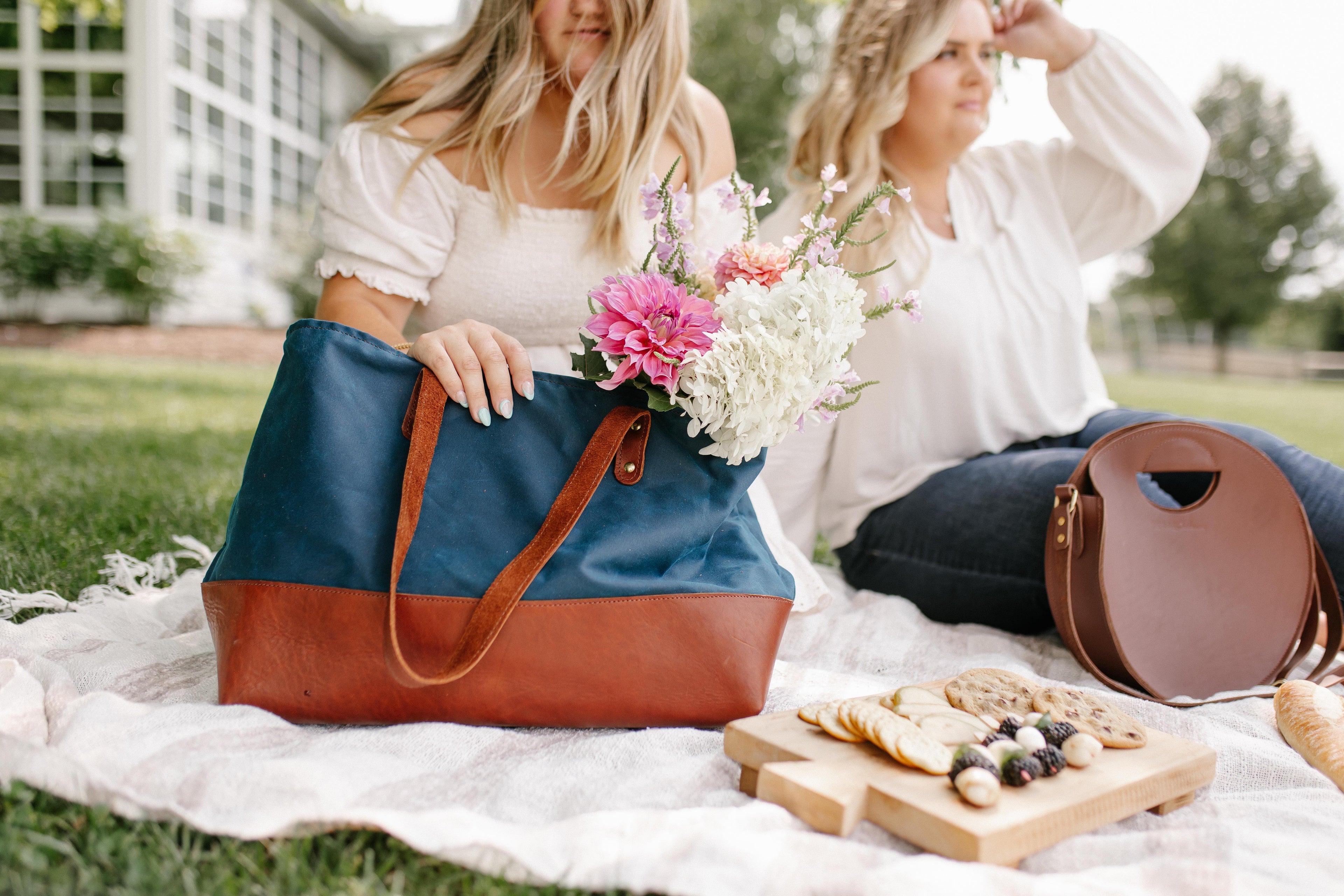 Two stylish women enjoy a blissful picnic with flowers, artisan cheese board, and fashionable tote bags on a sunny day outdoors.

