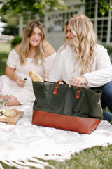 Two women enjoy a picnic with a green and brown leather tote bag, featuring fresh bread, cheese, and crackers, on a white blanket outdoors.
