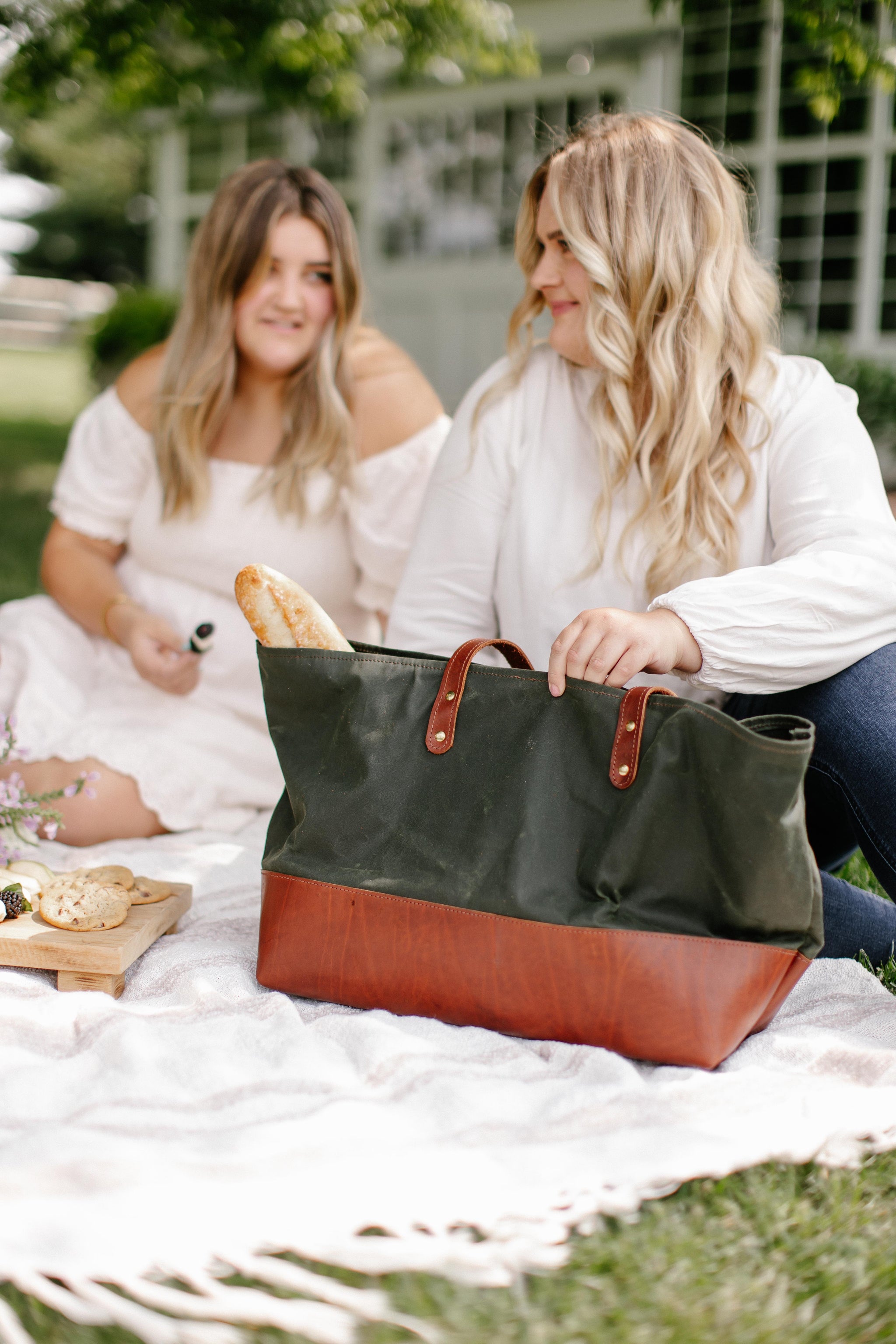 Two women enjoy a picnic with a green and brown leather tote bag, featuring fresh bread, cheese, and crackers, on a white blanket outdoors.
