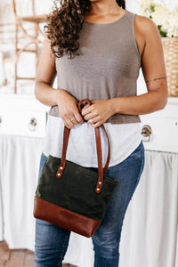 A woman models a stylish waxed canvas tote bag with leather accents in a bright, modern kitchen setting.

