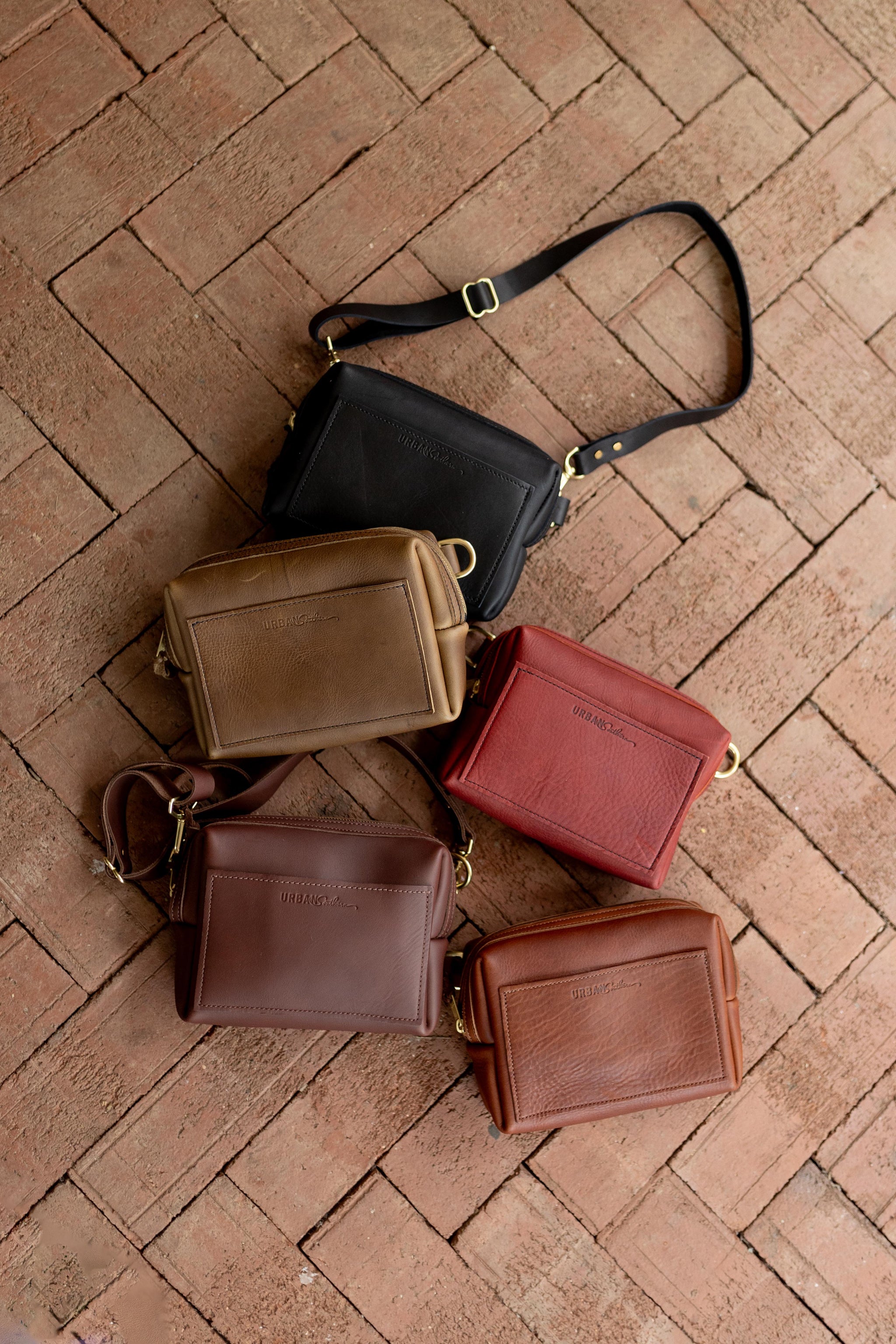 Displaying five Urban Artisan leather crossbody bags in shades of black, brown, and red on a brick background.
