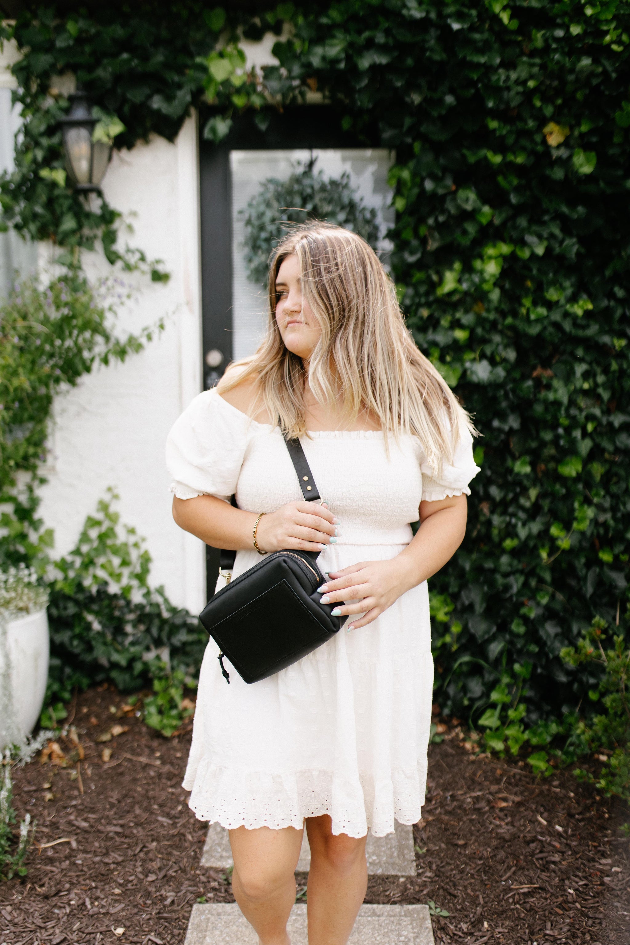 Woman models a white off-the-shoulder dress and black crossbody bag in front of an ivy-covered house.
