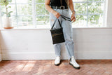Woman holds an Urban Originals black crossbody bag, wearing ripped jeans and white boots inside a bright room.
