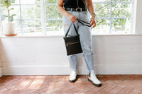 Woman holds an Urban Originals black crossbody bag, wearing ripped jeans and white boots inside a bright room.
