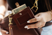 Woman holds a brown leather wallet with a dollar bill sticking out near a matching purse.
