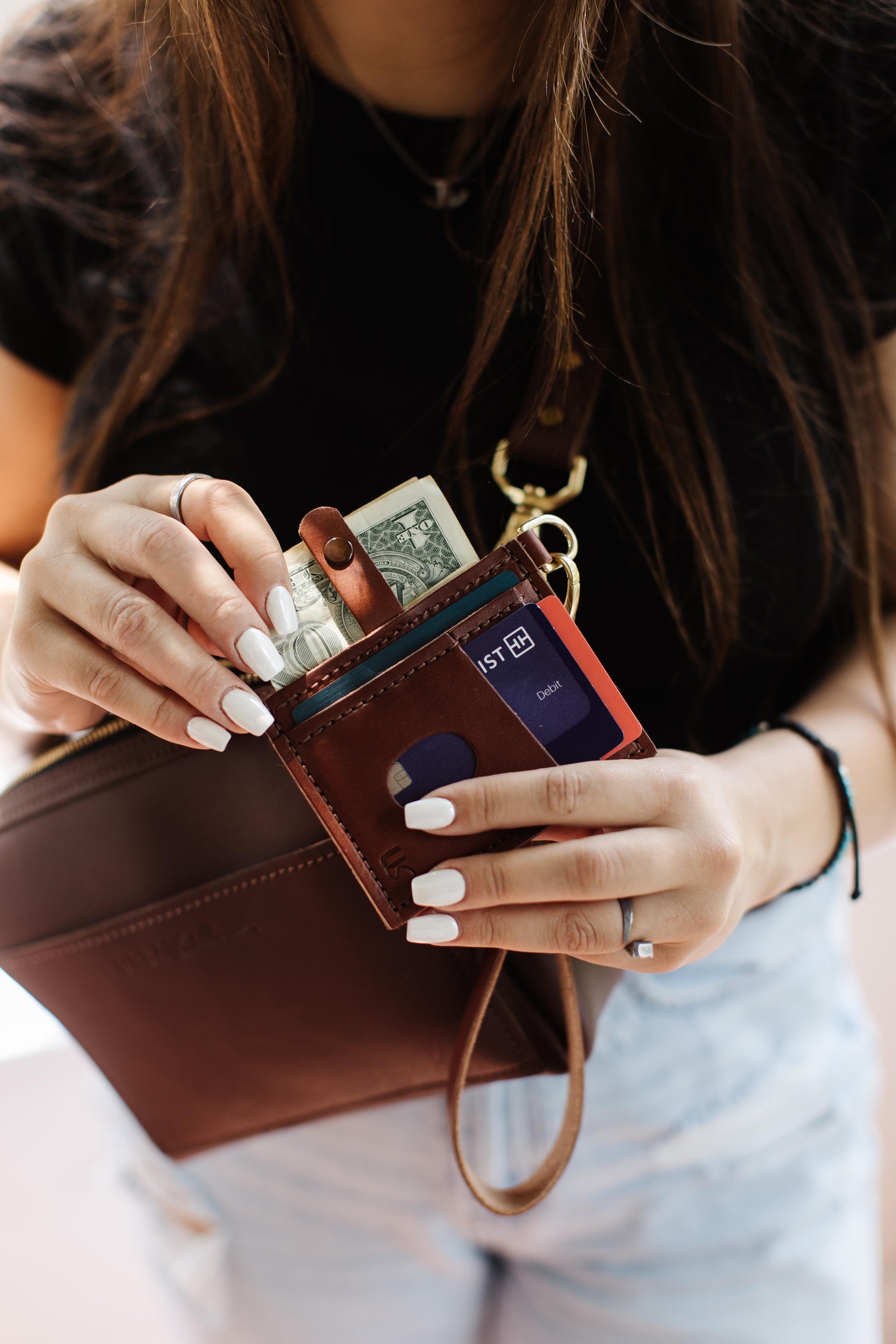 Woman pulls out cash and a debit card from a brown leather wallet attached to a crossbody bag.
