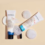 Mini Bloom's Knots Be Gone detangler and Fresh N' Clean hair and body wash sit amongst foamy sponges on a beige surface.
