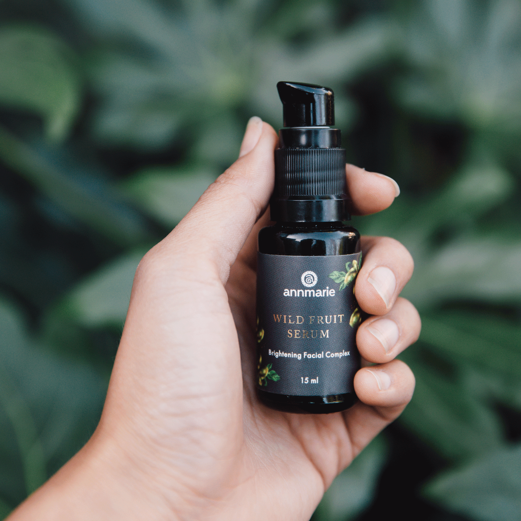 A hand holds Annmarie Wild Fruit Serum, a brightening facial complex, against a leafy green backdrop.
