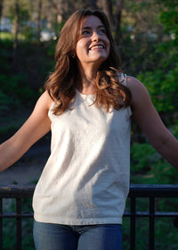 A young woman with brown hair smiles while looking up, standing by a park railing in the sunlight.
