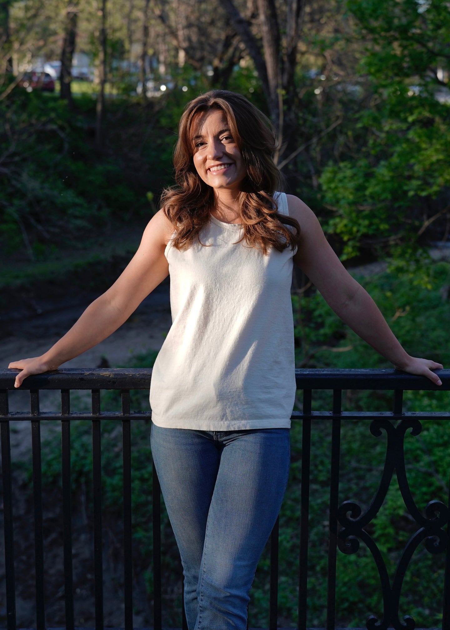 A woman smiles while leaning against an iron bridge railing in a park setting.
