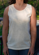 A woman models a simple, cream-colored organic cotton tank top and jeans outdoors.
