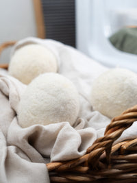 Three white wool dryer balls sit inside a rustic wicker laundry basket lined with soft linen fabric.
