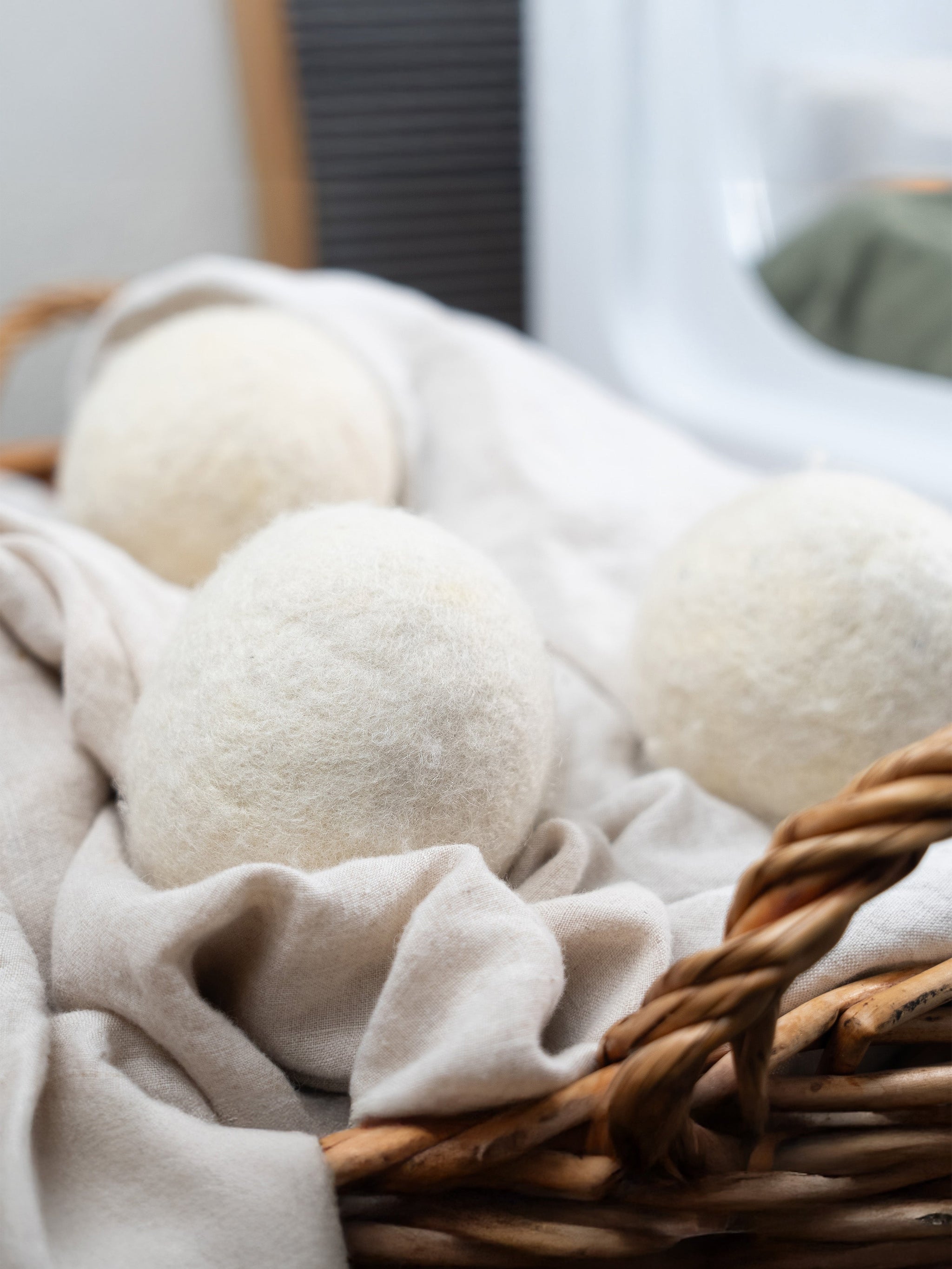 Three white wool dryer balls sit inside a rustic wicker laundry basket lined with soft linen fabric.
