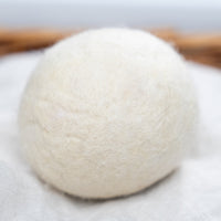 A white, fluffy wool dryer ball rests on a white linen cloth inside a brown wicker basket.
