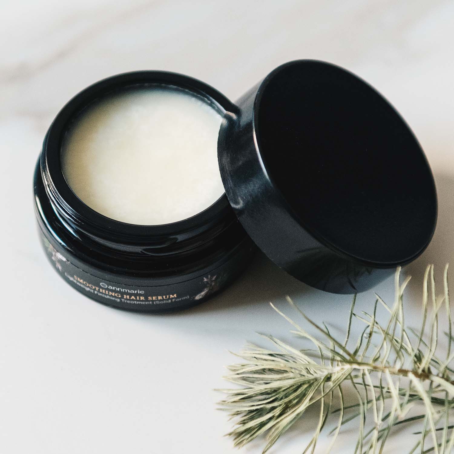Annmarie Smoothing Hair Serum sits open on a marble surface, its black lid resting beside a sprig of evergreen.
