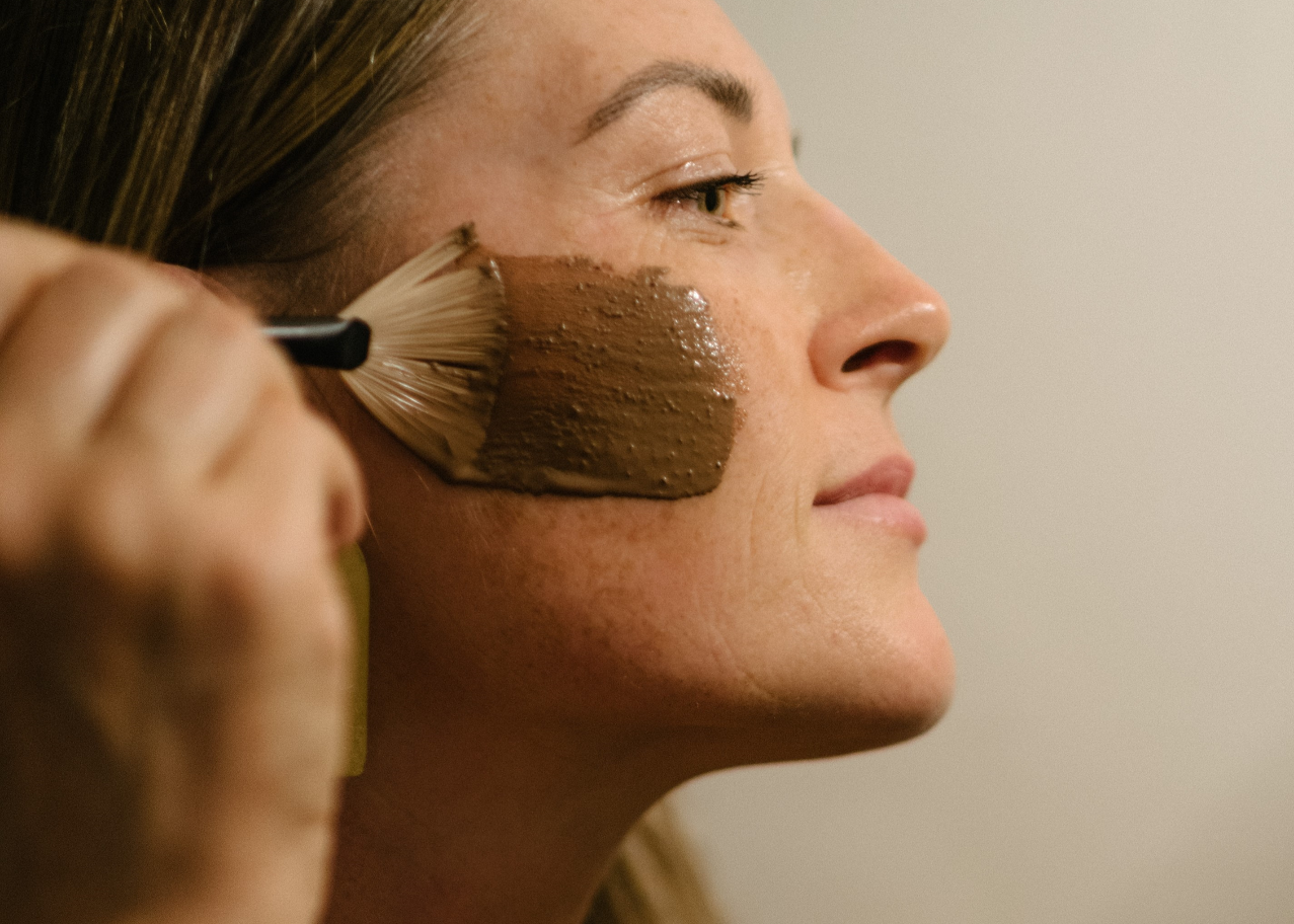 A close-up side profile of a woman applying a thick brown face mask or foundation with a brush against a plain light beige background.