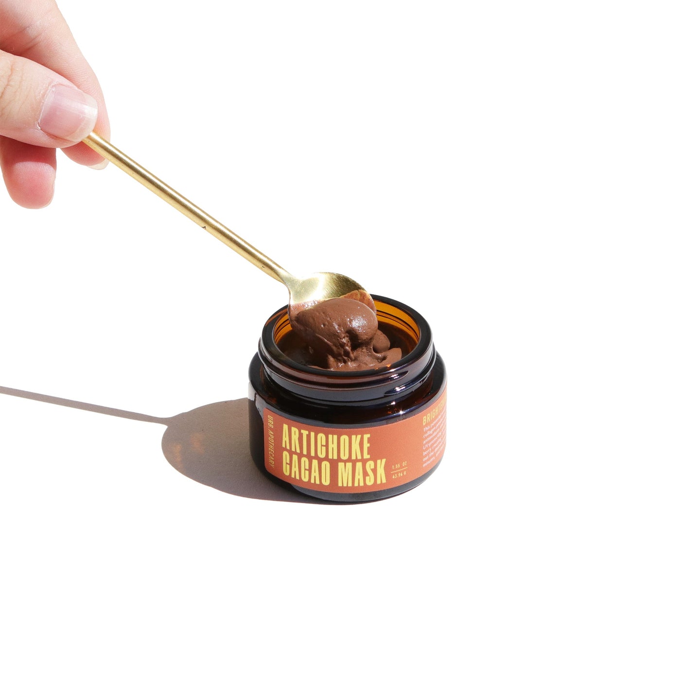 A hand scoops the Urb. Apothecary Artichoke Cacao Mask from its amber glass jar with a golden spoon.